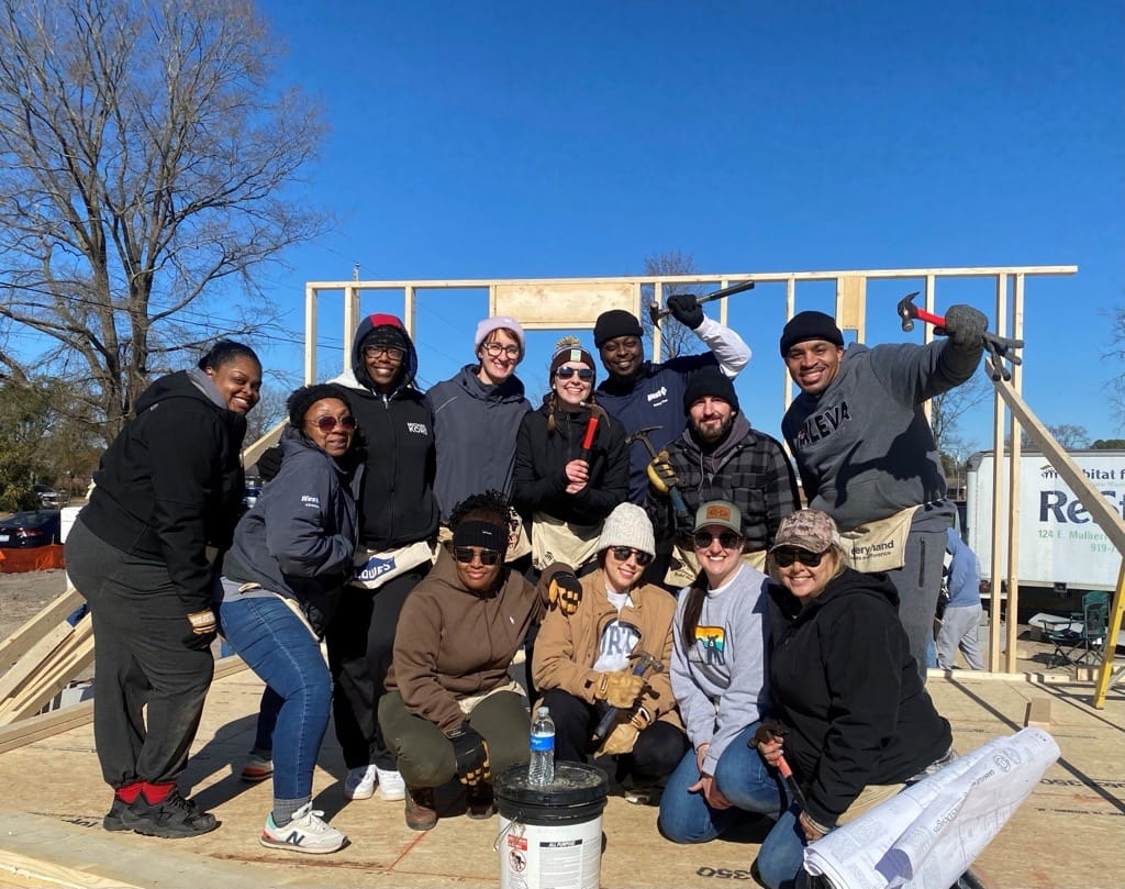 Employee volunteers posing at a Habitat for Humanity home construction site with a partially framed house in the background.